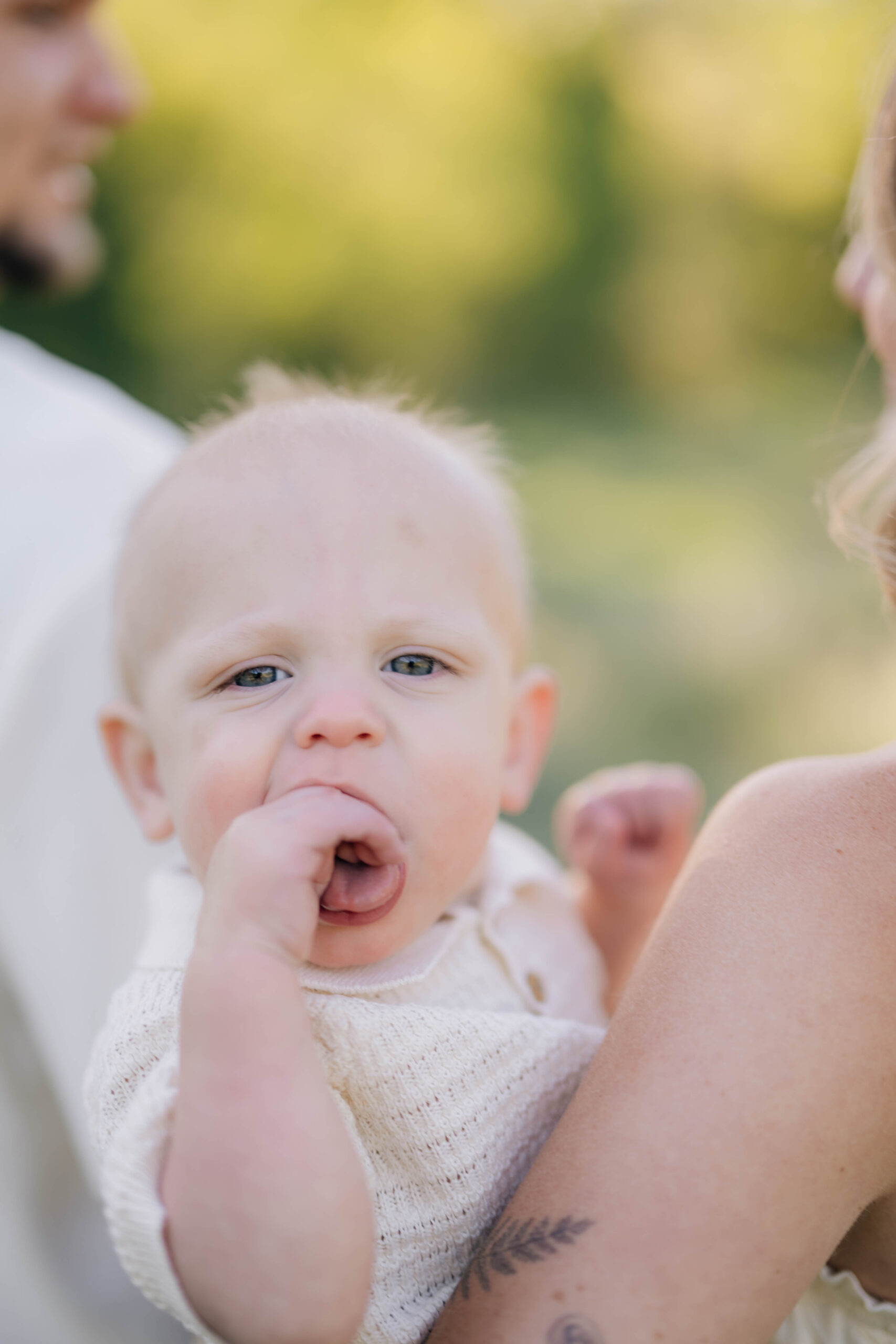 Close up of baby with his hand in mouth being held by mom. Parents are blurred in the back
